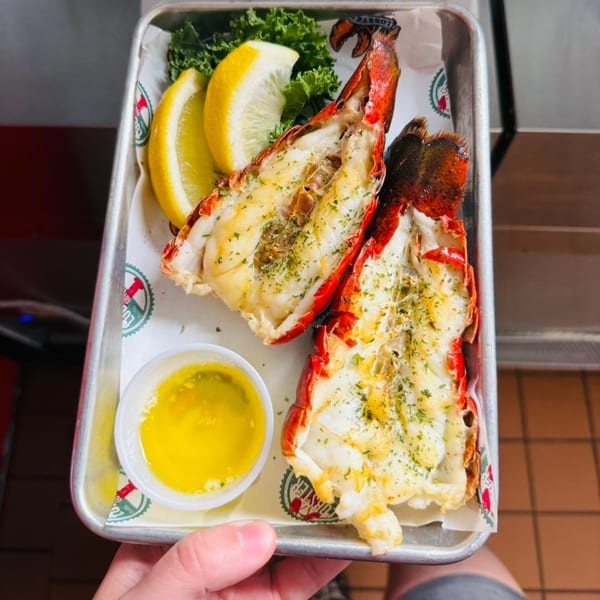Two grilled lobster tails with lemon wedges, parsley, and melted butter on a metal tray.