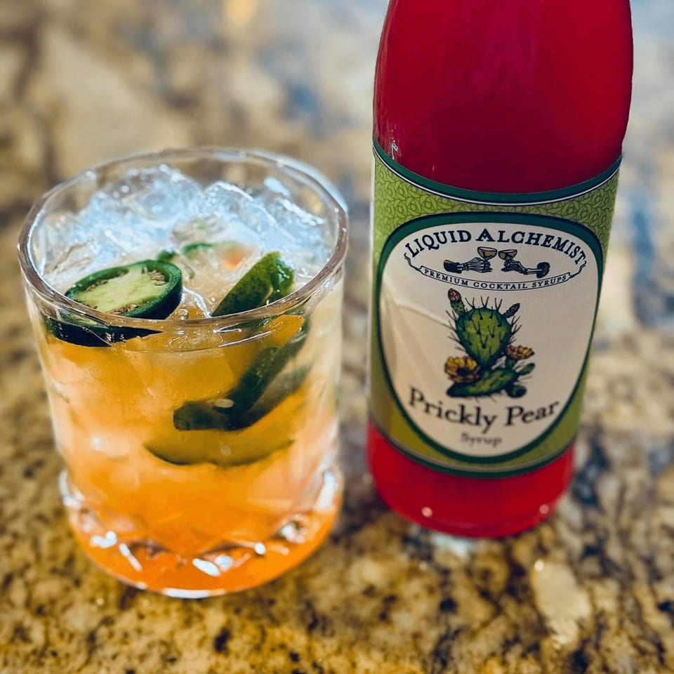 A cocktail with jalapeno slices and ice next to a bottle of Liquid Alchemist Prickly Pear Syrup.