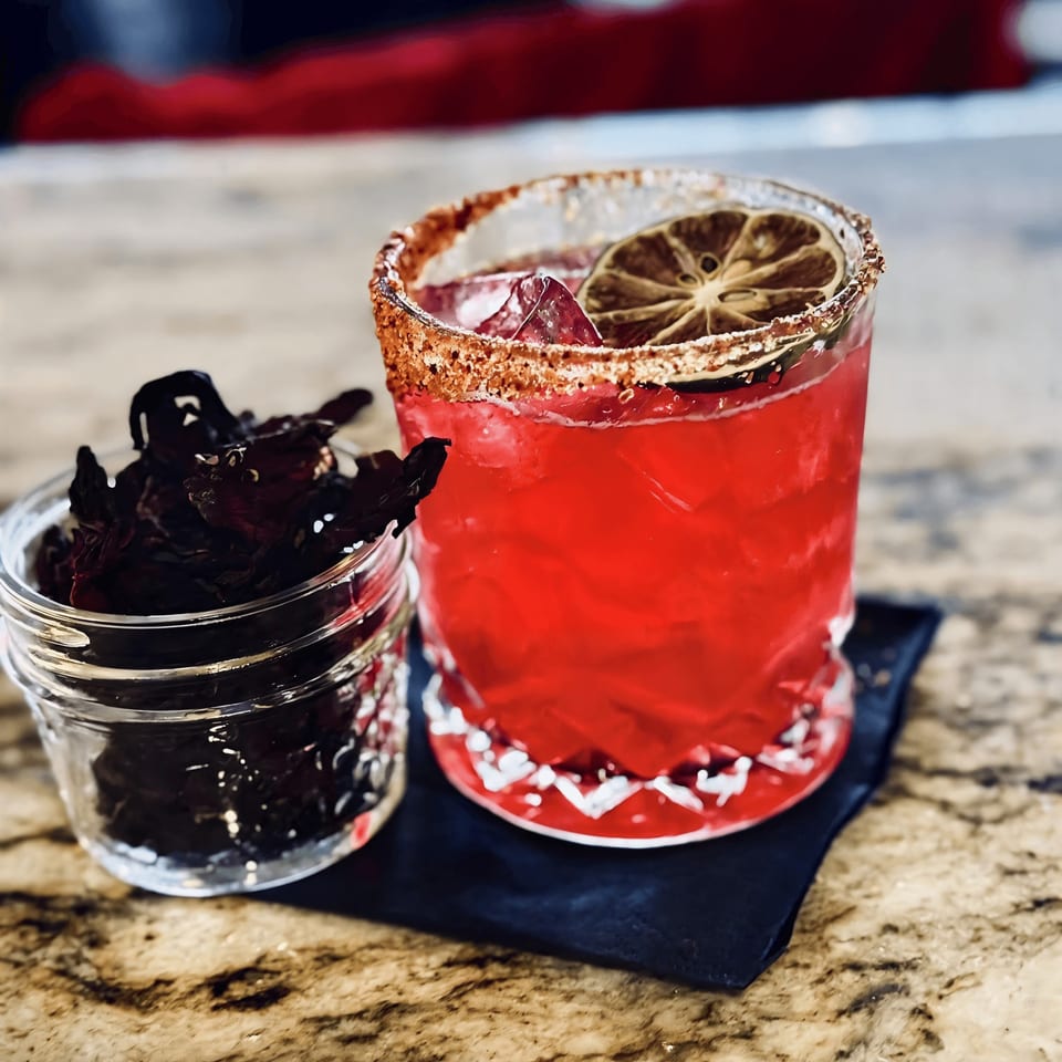 A vibrant red cocktail with a salted rim and lime garnish next to a jar of dried hibiscus.