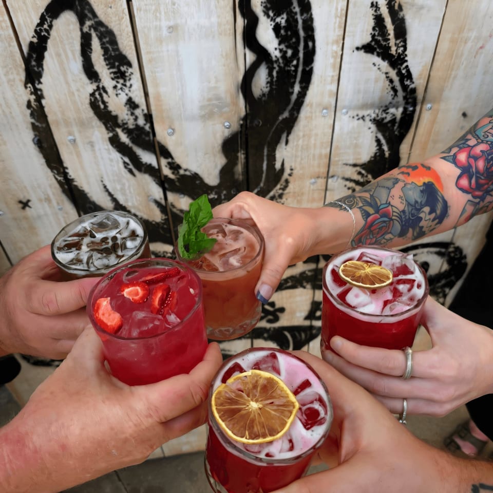 Five people toast colorful cocktails with ice and garnishes, against a wooden wall with a lobster graphic.