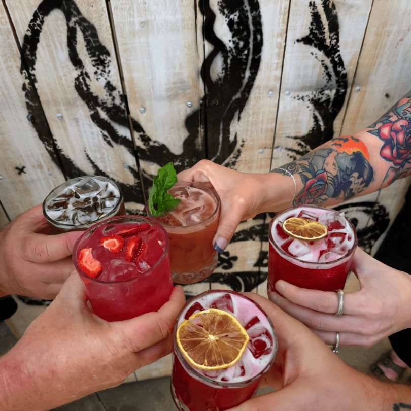 Five people toast colorful cocktails with ice and garnishes, against a wooden wall with a lobster graphic.