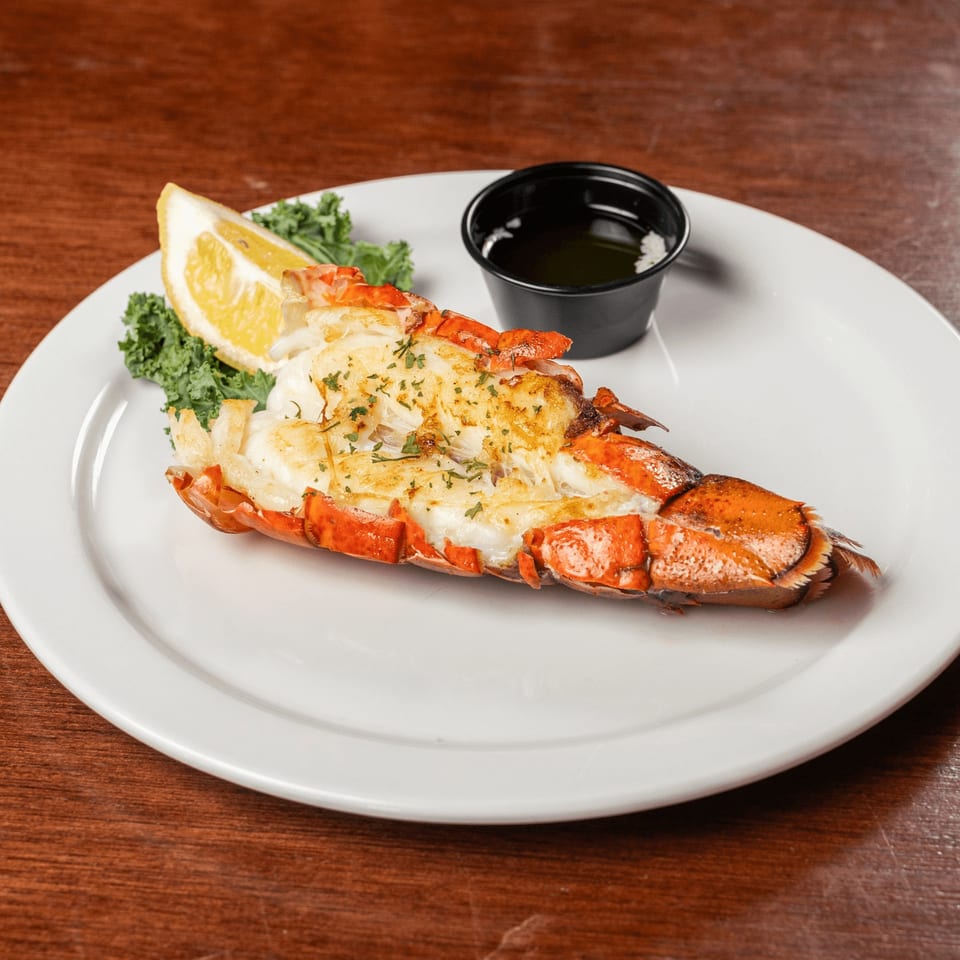 Grilled lobster tail with a lemon wedge, kale, and a side of butter on a white plate.